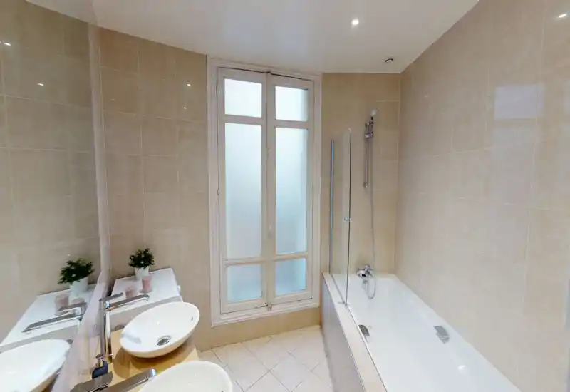 Salle de bains lumineuse et moderne avec baignoire/douche, fenêtre dépoli, double vasque sur plan et grand miroir mural ; carrelage beige neutre pour une atmosphère propre et spa-like.
