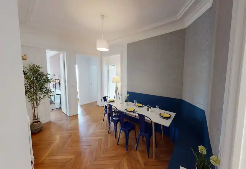 Coin repas lumineux et élégant avec une table blanche, des chaises métalliques bleues et une banquette intégrée devant un papier peint à motifs — dressé pour un repas, idéal pour partager.
