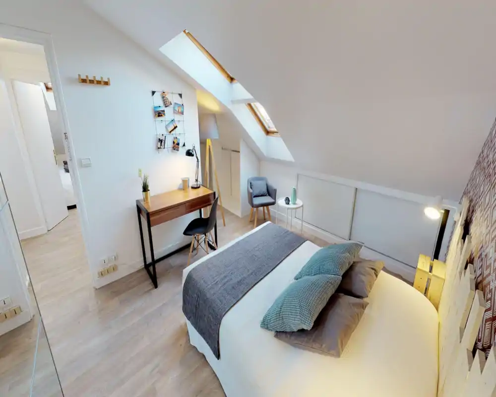 Chambre mansardée élégante avec un lit double confortable, oreillers moelleux et un jeté gris. Les fenêtres de toit apportent une belle lumière naturelle ; un petit bureau et un fauteuil offrent un coin travail/lecture agréable.