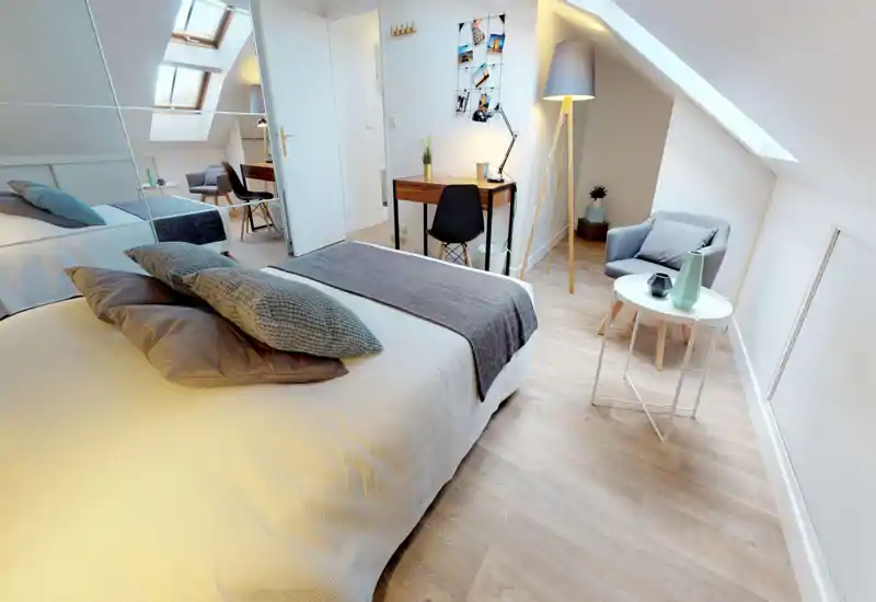 Chambre mansardée lumineuse et moderne avec un grand lit confortable, coussins texturés et un coin lecture près du velux. Un petit bureau et une lampe contemporaine complètent cet espace accueillant.