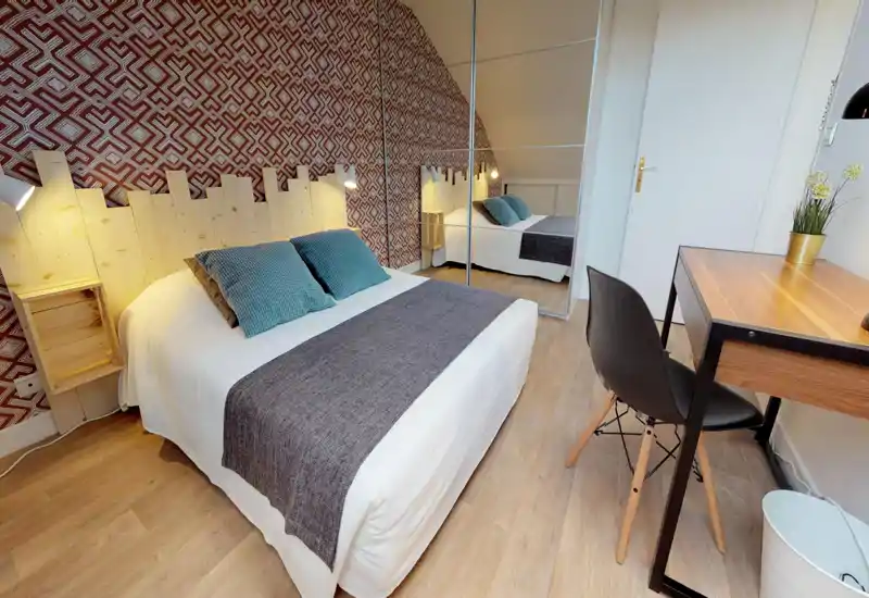 Chambre lumineuse et soignée avec un lit double confortable, coussins décoratifs et tête de lit en bois ; une armoire miroir agrandit l'espace et un petit bureau offre un coin travail.