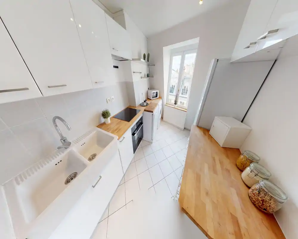 Cuisine moderne et lumineuse en enfilade avec placards blancs, plans de travail en bois, four encastré et évier double près de la fenêtre apportant une belle lumière naturelle.