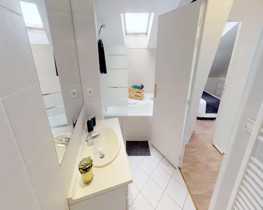 Salle de bains moderne et lumineuse avec lavabo, miroir et baignoire sous une fenêtre de toit ; carrelage blanc et accents bois visibles vers la chambre.