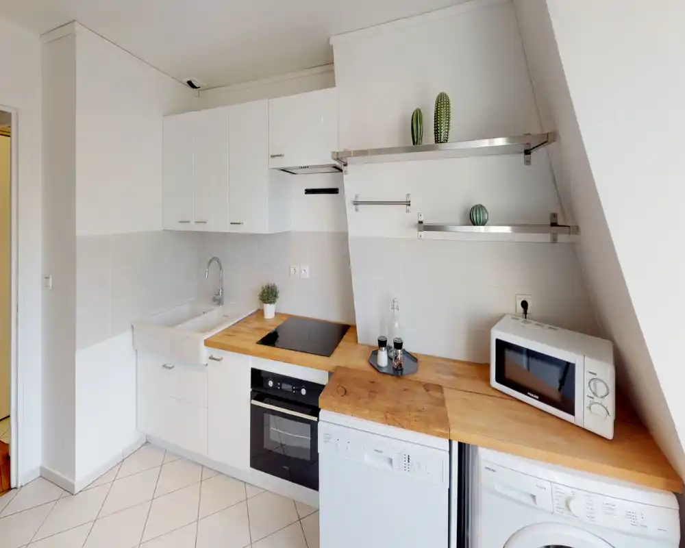 Cuisine compacte moderne et lumineuse avec meubles blancs, plans de travail en bois, four encastré et plaque à induction. Agencement fonctionnel avec évier, micro-ondes et lave-linge — propre et prêt à l'emploi.