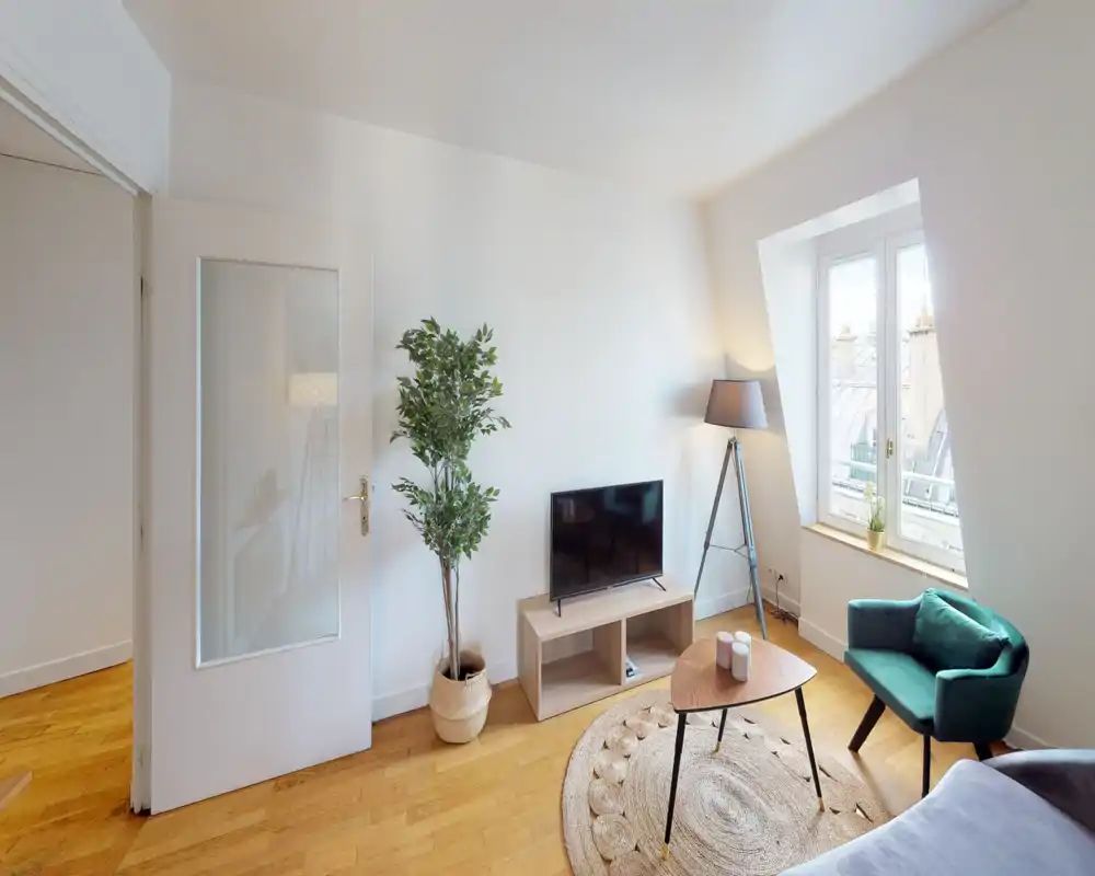 Salon lumineux et chaleureux avec meuble TV compact, fauteuil vert, tapis en fibres naturelles et grande fenêtre offrant une vue sur la ville — idéal pour se détendre ou recevoir.