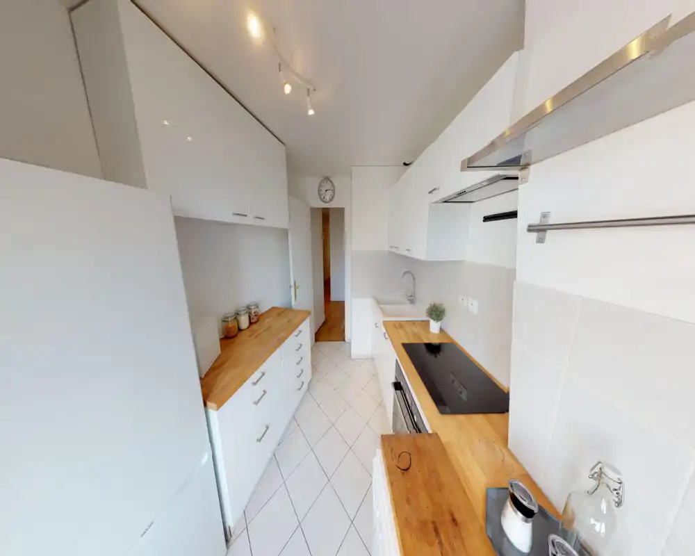 Cuisine moderne et lumineuse avec placards blancs, plans de travail en bois, plaque de cuisson et four intégrés ; sol carrelé et rangements pratiques, idéale pour petit appartement.