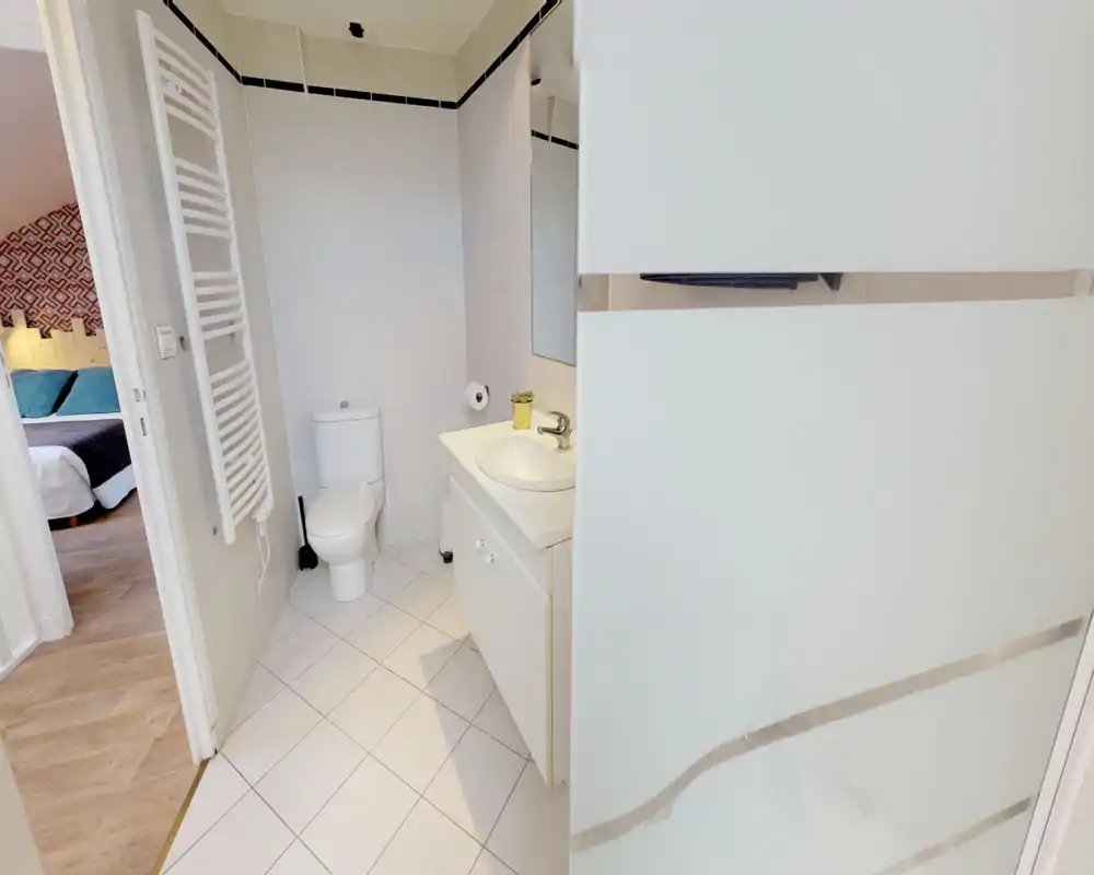 Salle de bain compacte et lumineuse avec WC, lavabo sur meuble et miroir ; carrelage blanc et radiateur sèche-serviettes pour une ambiance propre et moderne.