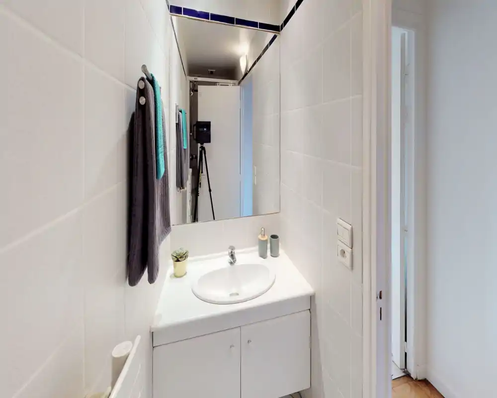 Salle de bain compacte et bien éclairée avec meuble vasque blanc, grand miroir et serviettes bien rangées — propre et moderne.