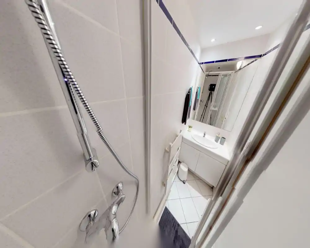 Salle de bains moderne et compacte avec carreaux blancs, vasque avec meuble, miroir et douchette au premier plan. Lumineuse, propre et fonctionnelle, idéale pour un petit appartement.