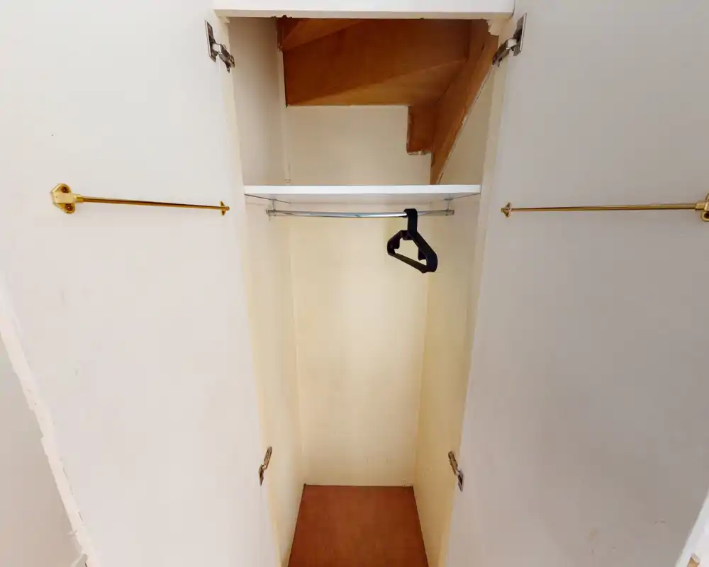 Pequeño armario empotrado con barra para colgar y un par de perchas de plástico; suelo con moqueta y balda bajo un techo inclinado.