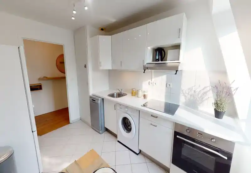 Bright modern kitchen with white cabinetry, built-in oven, induction cooktop and integrated washer/dishwasher. Clean countertops, jars and a potted plant add a welcoming touch.
