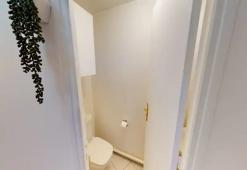 Small, clean toilet room with white tiles and neutral walls; compact fixture, wall-mounted toilet roll holder and a cabinet above the cistern.