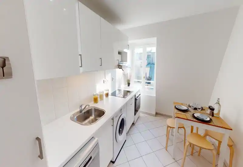 Bright compact kitchen with white cabinets, integrated appliances, a sink and a small wooden dining table set for four — ideal for a cozy city apartment.