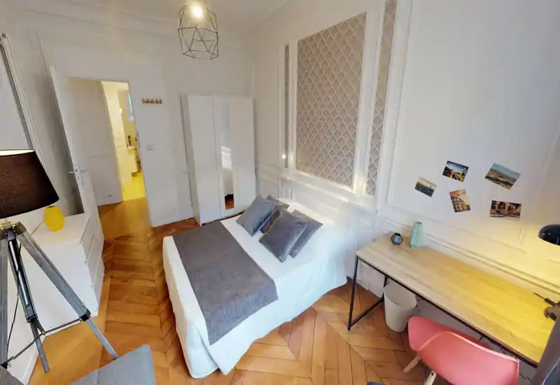 Bright, elegant bedroom with a centrally placed double bed, textured headboard panels, wooden herringbone floor and a small workspace by the wall — comfortable and stylish for short or long stays.