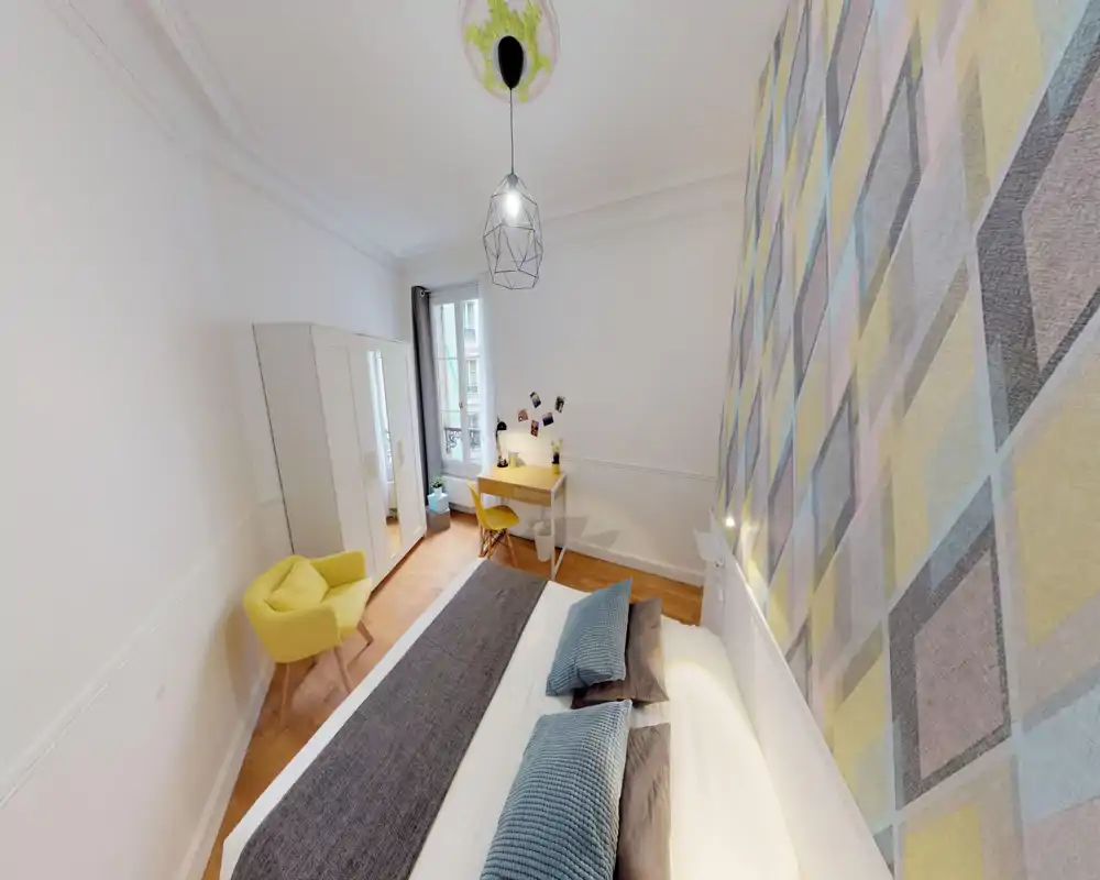 Chambre lumineuse et bien décorée avec un lit double confortable, coussins texturés et un mur à motif; on trouve une armoire, un bureau et un fauteuil jaune près de la fenêtre.