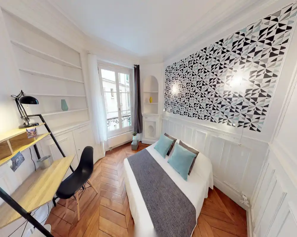 Chambre lumineuse et stylée avec lit double, mur à motif et lumière naturelle provenant d'une grande fenêtre. Parquet en chevrons chaleureux et petit espace de travail pour plus de confort et de fonctionnalité.