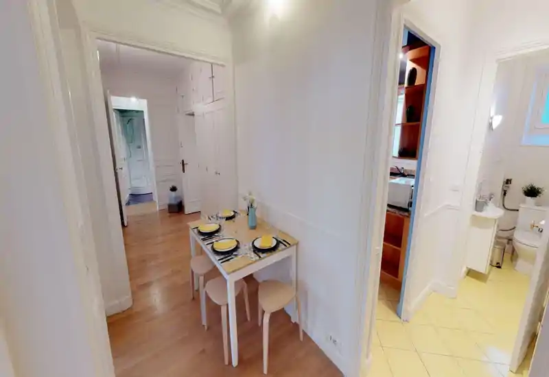 Compact dining area set for four in a bright hallway niche; simple wooden table and stools with colorful placemats create a neat, welcoming spot for meals.