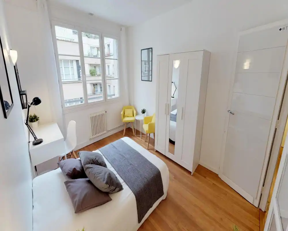 Chambre lumineuse et soignée avec un lit double en tons neutres, un bureau près de la fenêtre et deux fauteuils jaunes formant un coin détente chaleureux.