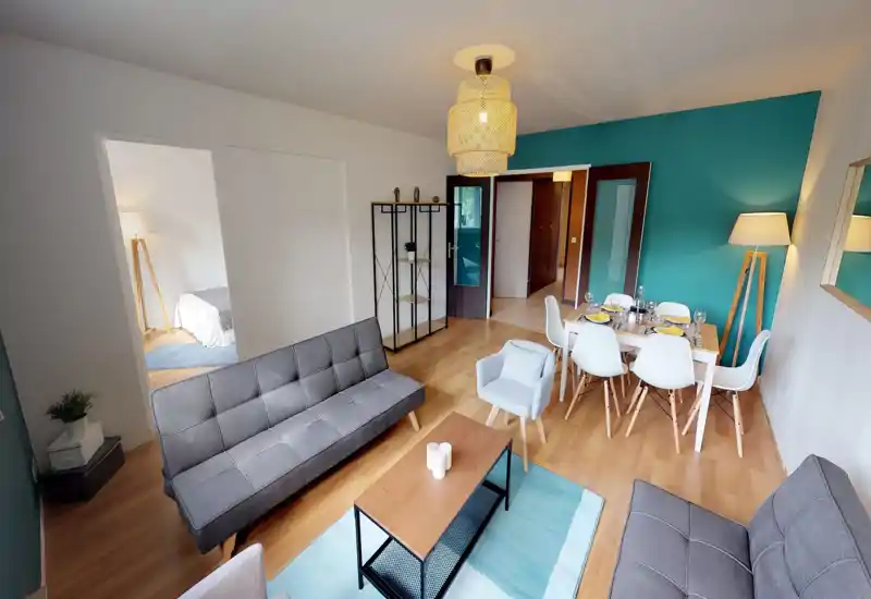 Spacious, modern living room with mid-century style sofas, a coffee table, and a dining area. Bright teal accent wall and warm wooden flooring create a welcoming atmosphere.