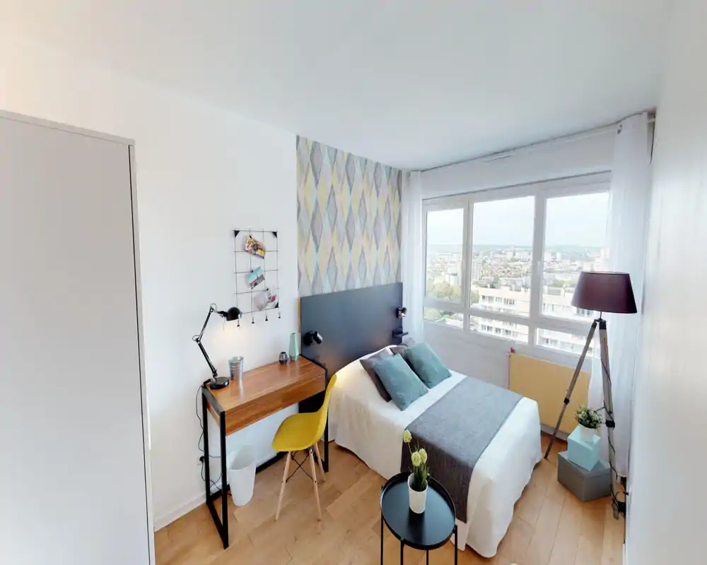 Chambre lumineuse et bien aménagée avec un lit confortable, coussins décoratifs et un bureau près de la fenêtre offrant une vue sur la ville. Grande fenêtre apportant une belle luminosité, mise en scène soignée pour location.