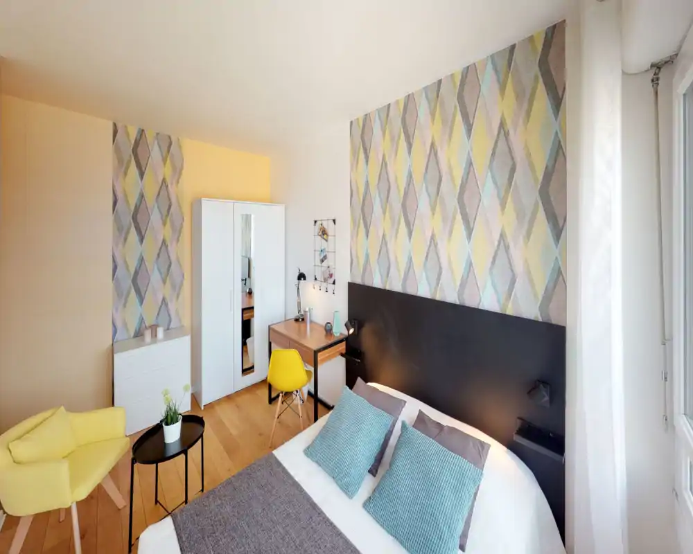 Chambre lumineuse et stylée avec un mur à motif, lit double confortable avec coussins et un coin bureau près de l'armoire. La lumière naturelle, le parquet chaud et le fauteuil jaune créent une ambiance accueillante.
