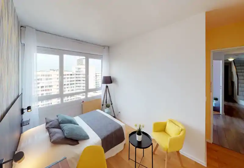 Sunny, modern bedroom with a double bed positioned by large windows that offer city views. The room features soft textiles, a standing lamp and bright yellow accent chairs for a cozy, contemporary feel.