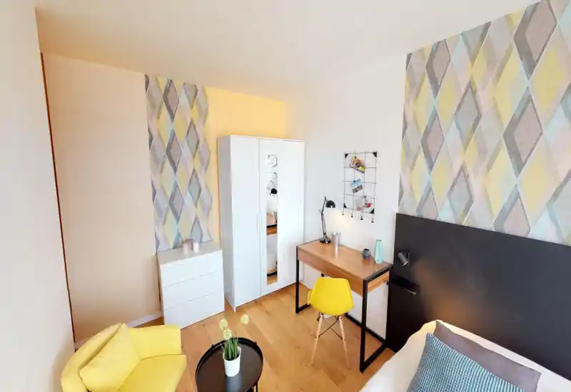 Bright, modern bedroom with a double bed framed by a dark headboard, patterned wallpaper, a small work desk and a yellow accent chair — cozy and well styled for rental listings.