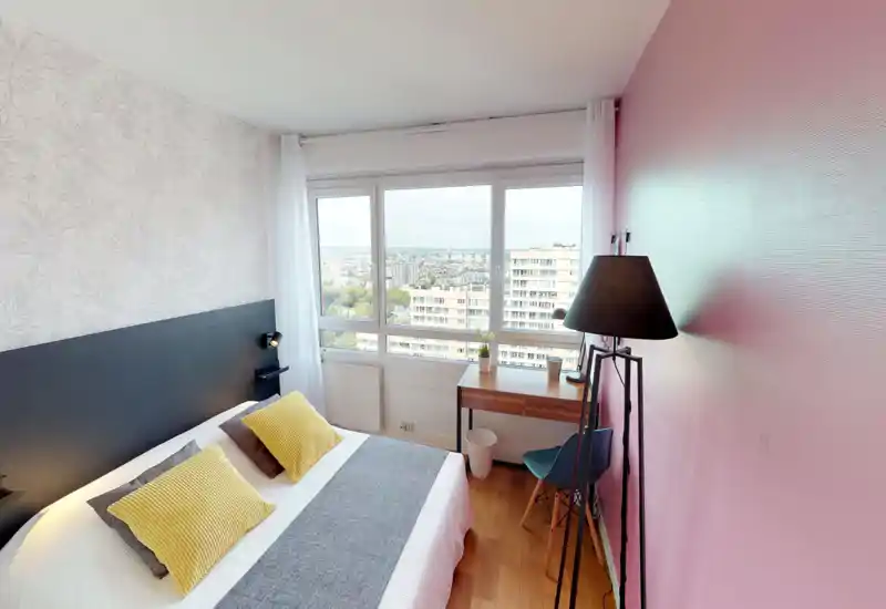 Bright, well-styled bedroom with a double bed dressed in neutral linens and yellow accent pillows. Large window offers city views and fills the room with natural light; small desk and floor lamp create a cozy work corner.