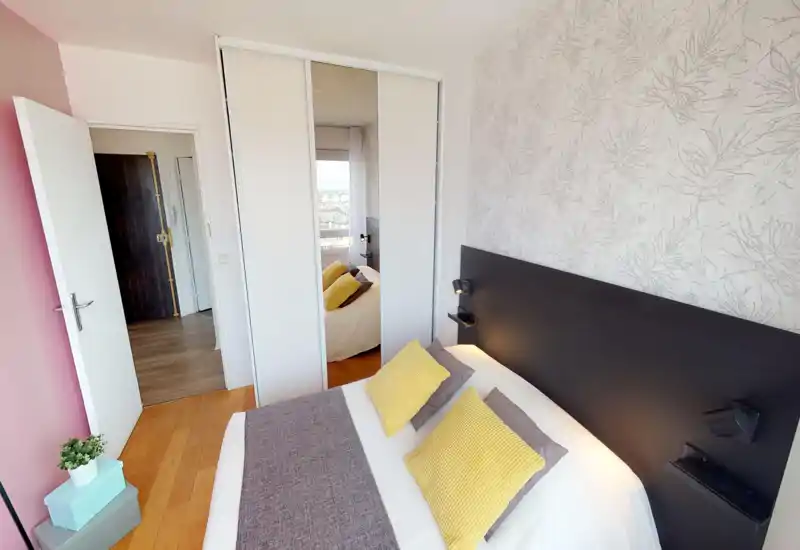 Bright, modern bedroom with a double bed dressed in coordinated cushions and throws, a dark headboard, and a mirrored wardrobe. Warm wooden floor and soft decorative wallpaper complete the cozy, well-lit space.
