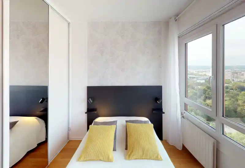 Bright, modern bedroom with a centered double bed, yellow accent cushions, built-in mirrored wardrobe and large windows offering city and park views. The room is neatly staged with a minimalist headboard and bedside reading lights.