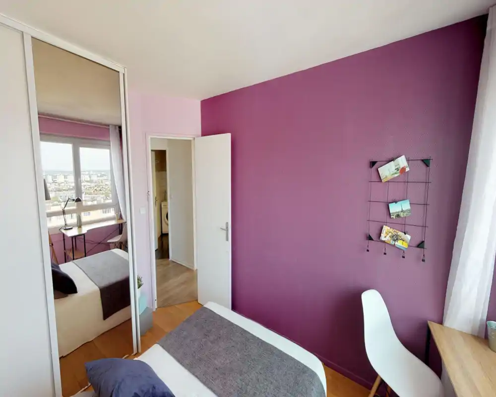 Camera da letto luminosa e arredata con letto matrimoniale, tessili accoglienti e una scrivania vicino alla finestra con vista sulla città. La parete color porpora e l'armadio con ante a specchio aggiungono carattere e profondità.