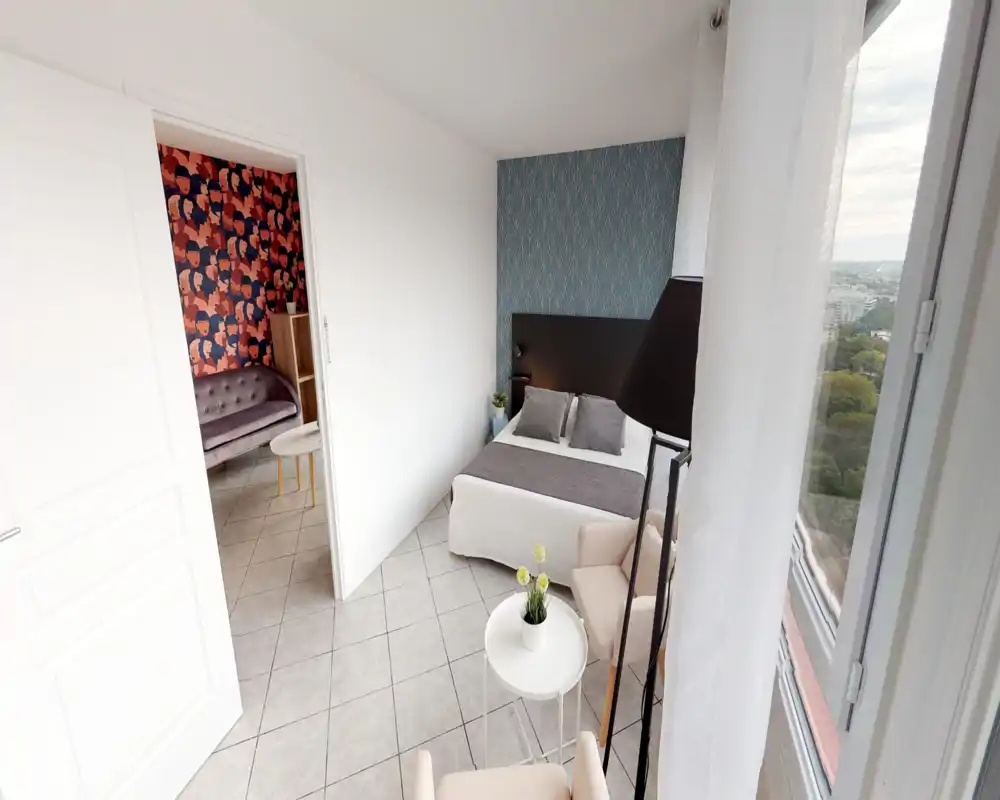 Chambre double lumineuse et bien mise en scène avec un mur décoratif, une tête de lit et une grande fenêtre apportant lumière naturelle et vue sur la ville. Linge neutre, coussins et coin salon créent une atmosphère chaleureuse et moderne idéale pour une annonce.