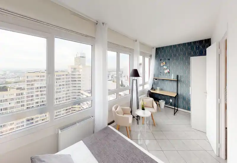 Chambre lumineuse et moderne avec de grandes fenêtres panoramiques, lit bien fait au premier plan et un coin détente avec table et fauteuils près de la fenêtre. Mur accent décoratif et petit bureau pratique pour travailler ou se détendre.