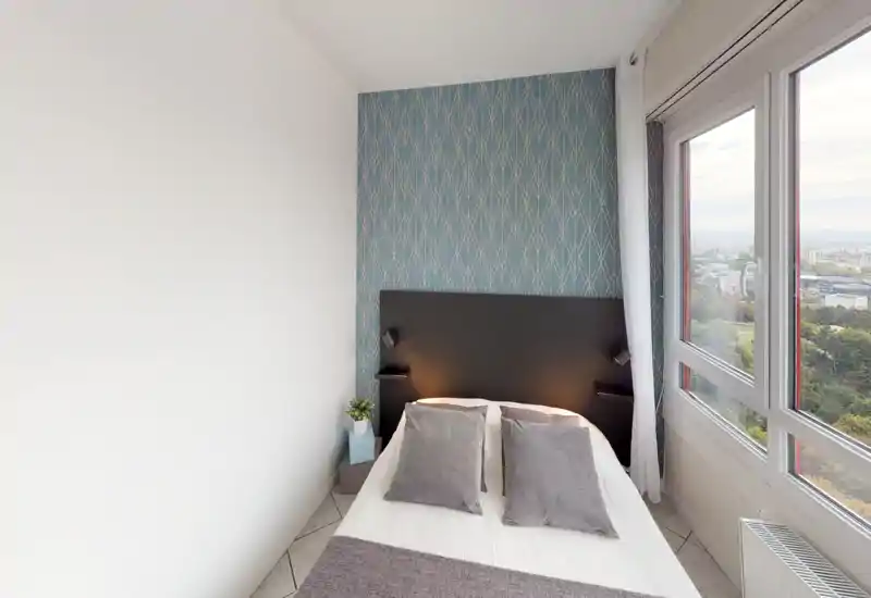 Chambre compacte et lumineuse avec lit double, papier peint accentué et grandes fenêtres offrant une vue sur la ville. Lampes de chevet douces et coussins décoratifs apportent une ambiance chaleureuse et accueillante.