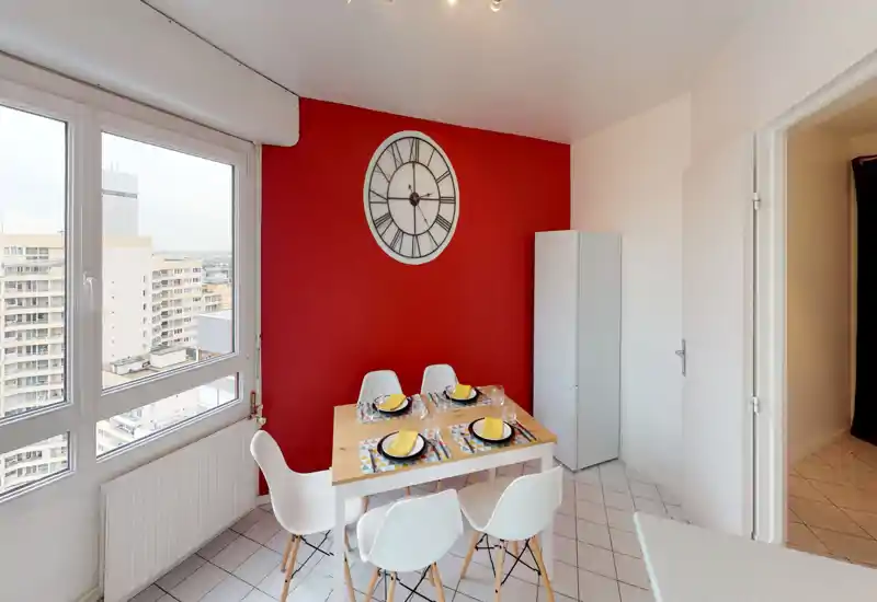 Bright dining corner with a wooden table set for four, modern white chairs and a bold red accent wall featuring a large decorative clock. Large windows provide city views and natural light, creating an inviting space for meals.