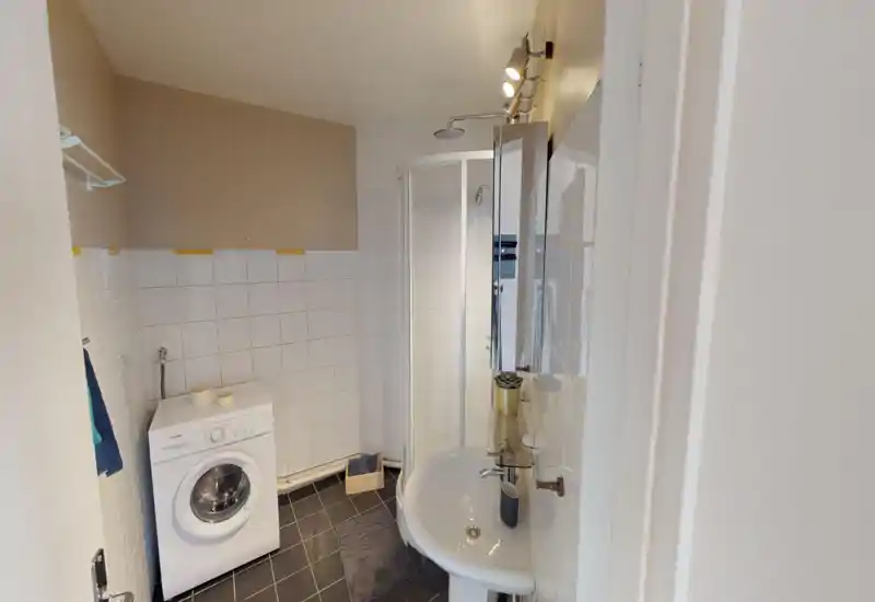 Compact bathroom with a corner shower cabin, small sink, and a front-loading washing machine; tiled walls and dark floor tiles create a clean, practical space.