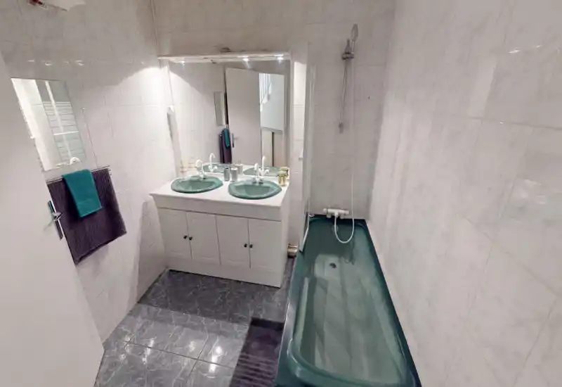 Bright, clean bathroom with a long teal bathtub, dual sinks set in a white vanity, and wall-mounted shower head. Polished tile walls and floor provide a neat, modern look ideal for rental listings.