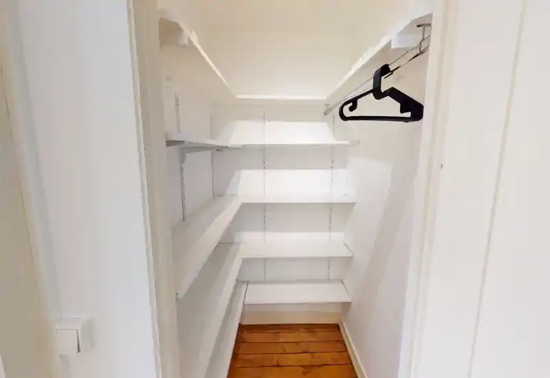 Small walk-in closet with white built-in shelving and a hanging rail, wooden floor and bright neutral walls — ideal for organized storage.