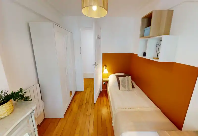 Compact single bedroom with a neat bed against a warm terracotta accent wall, bedside lamp and floating shelving; bright natural light and clean wooden floors create a cozy, well-styled space.