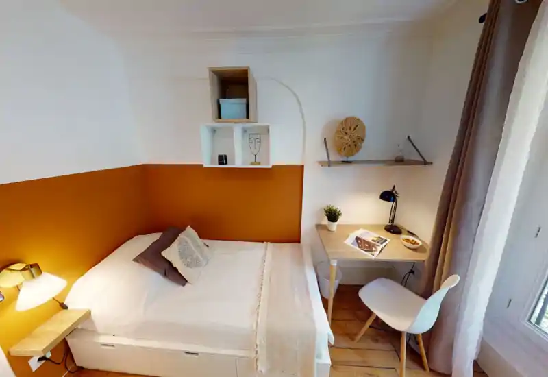 Bright, cozy bedroom with a double bed, warm ochre accent wall, bedside lamp and a compact workspace by the window.