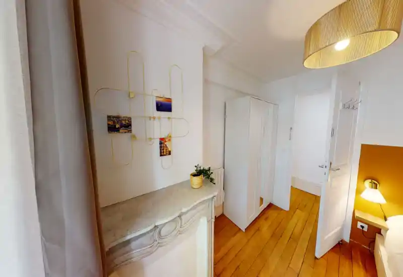 Compact bedroom corner with a warm wooden floor, bedside lamp and a partial view of the bed. The space is styled with soft textiles, a decorative chest and ambient lighting for a cozy feel.