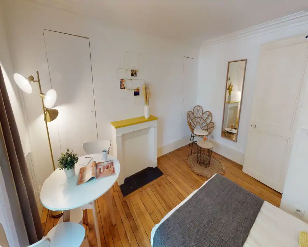 Chambre lumineuse et aménagée avec coin de lit visible, parquet, petite table ronde et chaise décorative ; décoration chaleureuse et moderne, idéale pour une annonce.