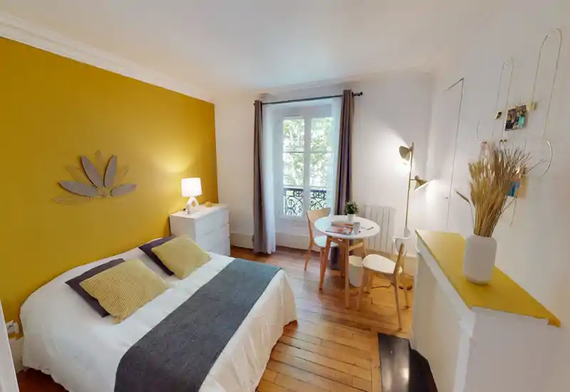 Bright, stylish bedroom with a mustard accent wall, a double bed dressed in white linens and coordinated cushions, and a small dining/study table by the window. Hardwood floors and tasteful lighting create a warm, inviting atmosphere ideal for short-term rentals.