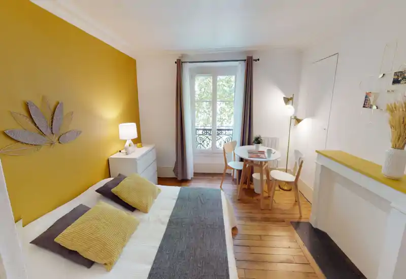 Sunlit, styled bedroom with a mustard accent wall, comfortable double bed dressed with textured cushions and a neat bedside table. The room features warm wooden floors, a small dining table by the window and modern decorative touches for a cozy, contemporary feel.