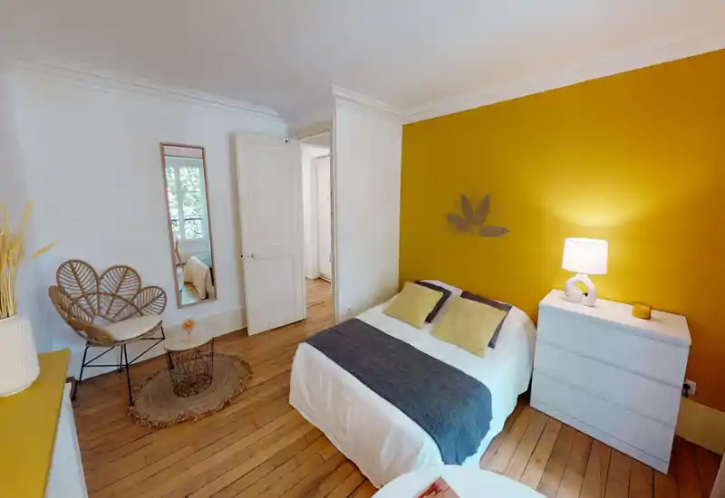 Well-styled bedroom with a mustard accent wall, double bed dressed in neutral linens and contrasting cushions, a bedside chest with lamp, and natural wood floors creating a warm, inviting atmosphere.