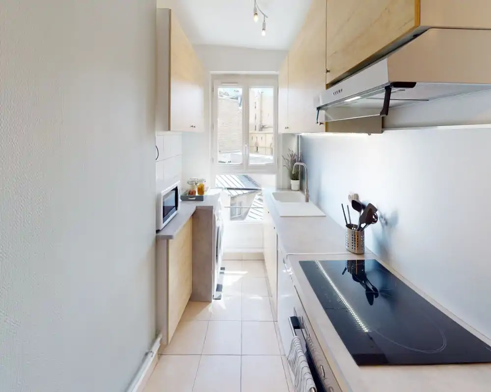 Cuisine moderne et étroite, lumineuse, avec placards en bois clair, plaque à induction, évier sous la fenêtre et appareils compacts — bien éclairée et optimisée pour un appartement citadin.