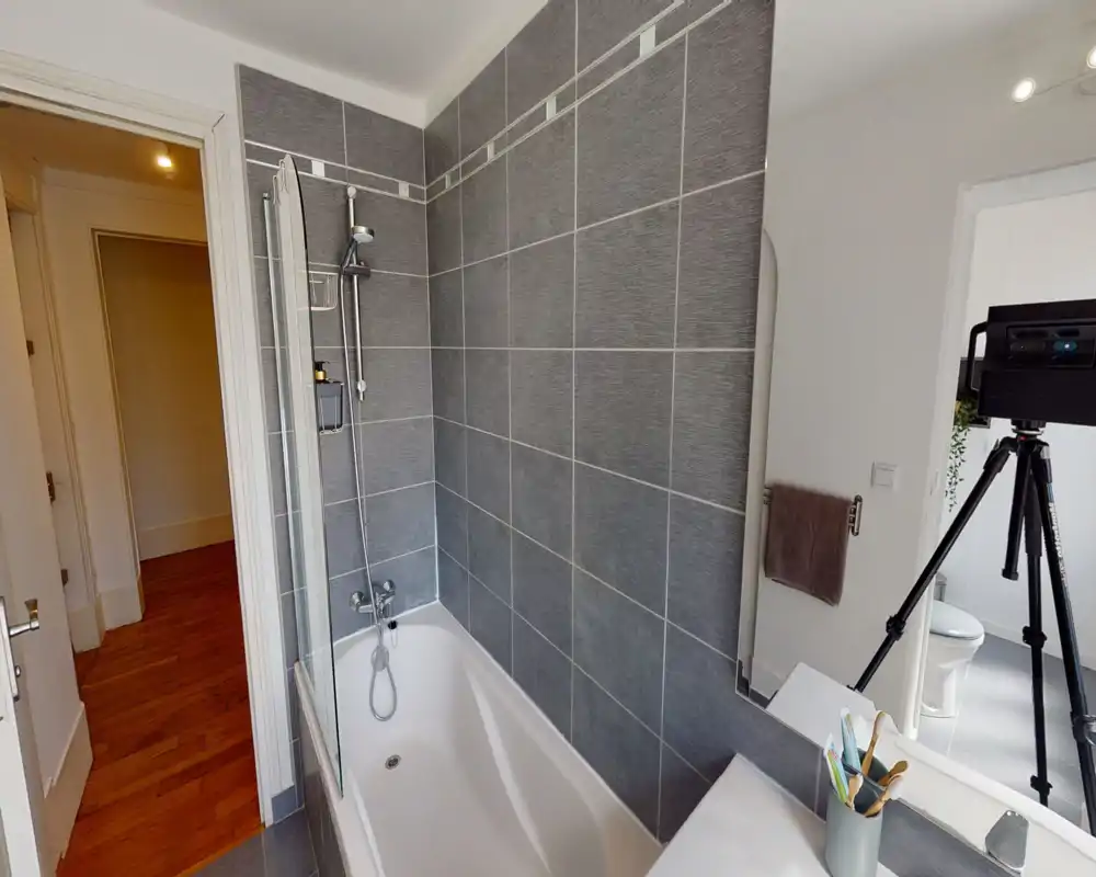 Salle de bains moderne avec combinaison baignoire-douche carrelée, douche murale et pare-douche en verre ; lavabo et miroir visibles avec accessoires rangés.