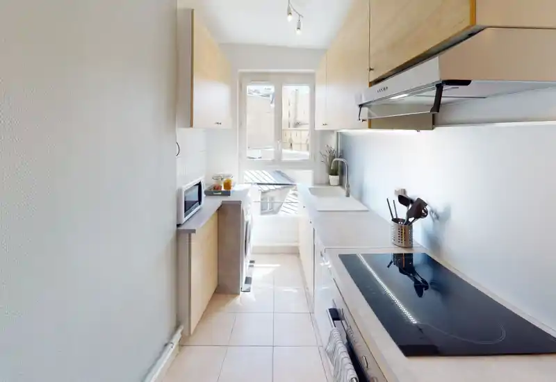 Bright, narrow modern kitchen with light wood cabinets, induction cooktop, sink under a window and compact appliances — well lit and efficiently laid out for city apartments.