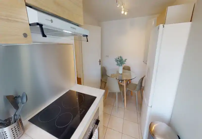 Compact modern kitchen with a glass-top dining table, four chairs and a refrigerator. Neutral tones, wood cabinetry and a cooktop create a bright, efficient cooking and eating area ideal for small apartments.