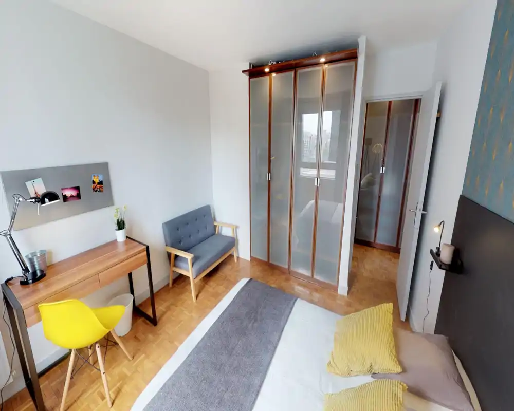 Chambre moderne et lumineuse avec un lit double confortable, coussins jaunes et un bureau en bois avec une chaise jaune. La pièce comprend un petit canapé gris, parquet et une armoire à portes dépoli, offrant un espace soigné et accueillant.
